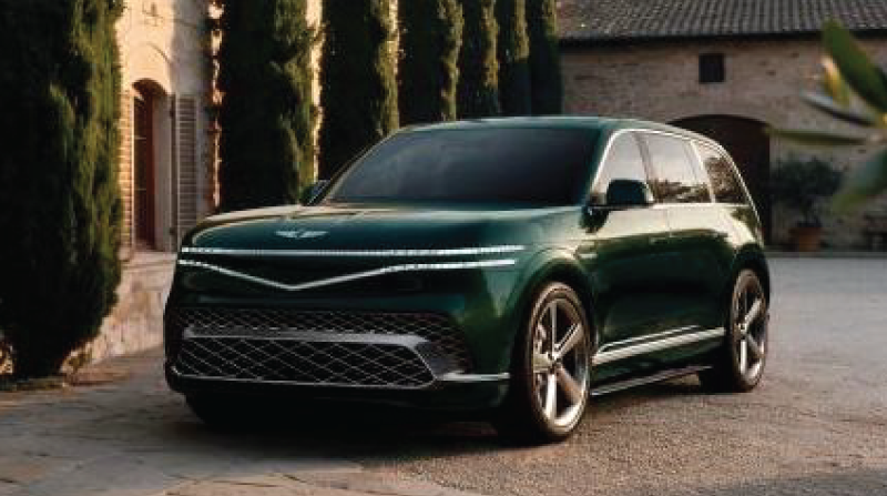 Dark green modern SUV with sleek, horizontal headlights parked on stone paving in front of a stone building and tall cypress trees.