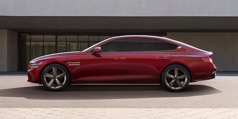 Red luxury sedan shown in side profile, parked in front of a modern building with large glass windows and a minimalist gray facade