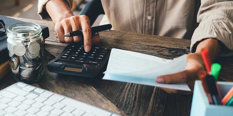 Person using a calculator while reviewing paperwork at a desk, with a jar of coins