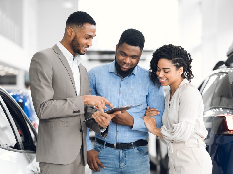 Three people standing in a car showroom review information on a tablet beside displayed vehicles