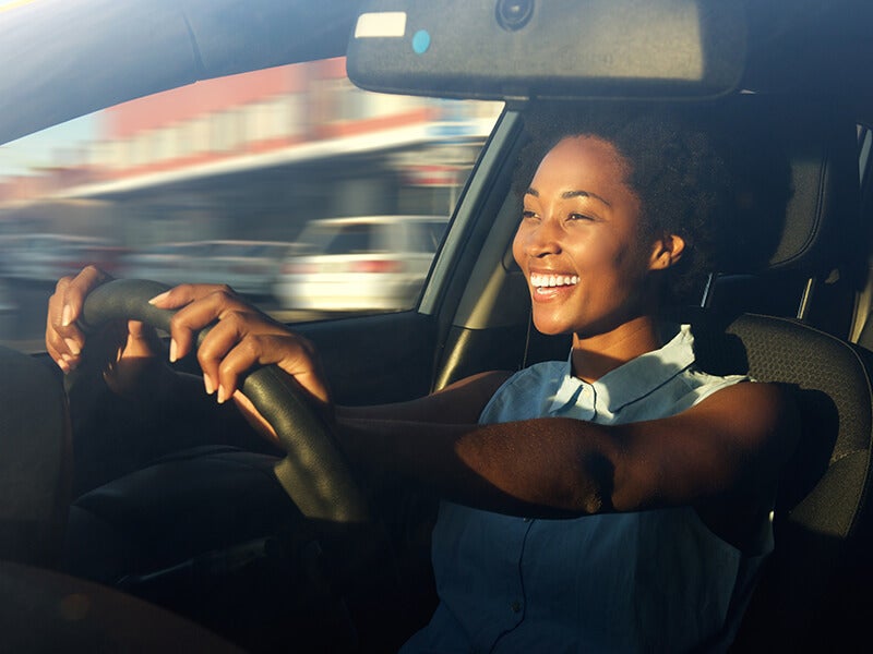 Person driving a car, shown from inside the vehicle, with hands on the steering wheel and sunlight illuminating the cabin