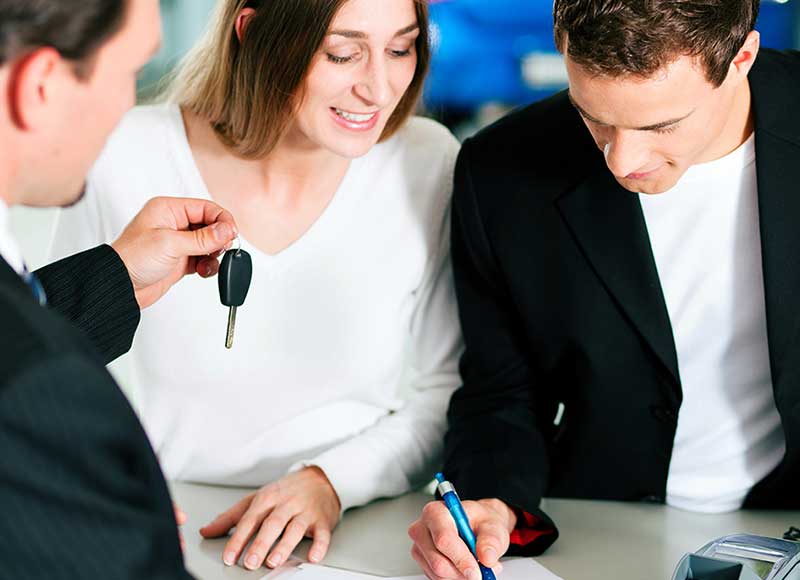 Person signing vehicle purchase paperwork while another person holds a car key nearby at a dealership desk