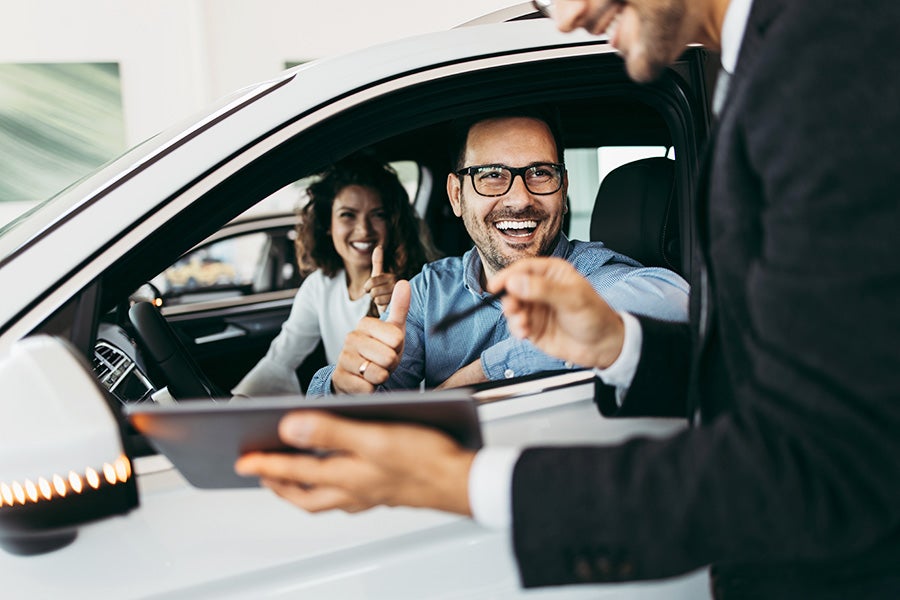 Customer seated in a white vehicle gives a thumbs-up while a dealership representative holds a tablet outside the driver-side window